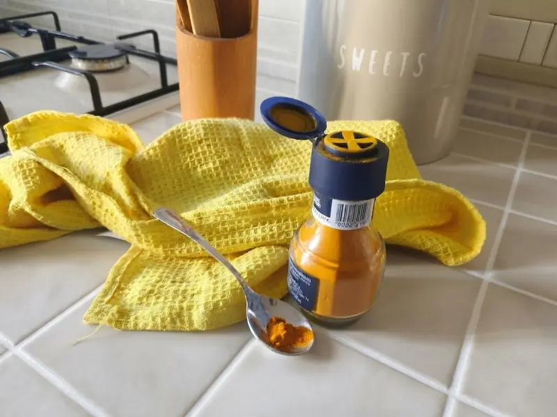 barattolo di curcuma su un ripiano di cucina piastrellato, accanto un cucchiaino con un pizzico di curcuma per condire il minestrone