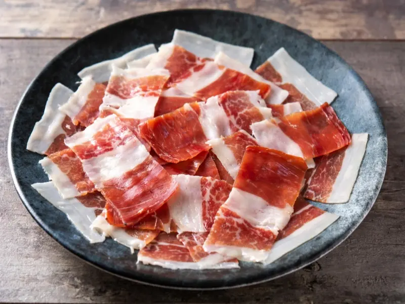 Fette di speck affumicato disposte su un piatto di ceramica scuro, pronte per essere usate nella lasagna di zucca e speck.