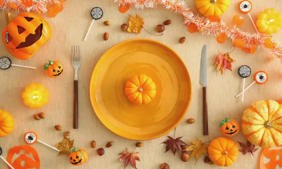 Allestimento di una tavola autunnale a tema Halloween vista dall'alto, con piatto, posate e decorazioni di zucche e foglie.